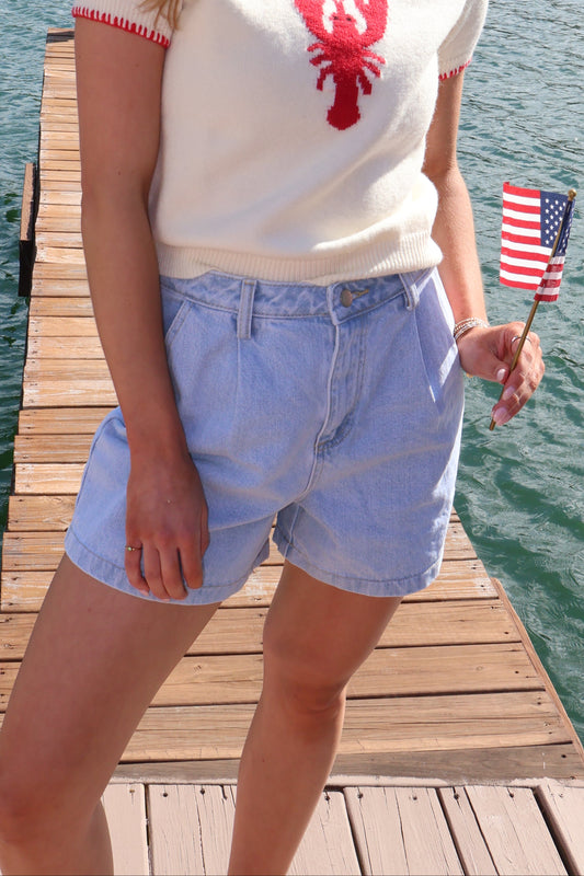 Woman standing on a dock by a lake, holding an American flag, wearing a lobster-themed shirt.